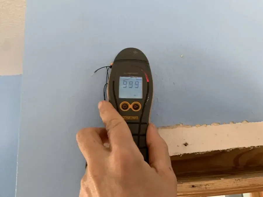 Handheld moisture meter pressed against a wall above a removed baseboard