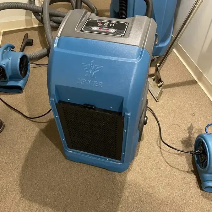 A dehumidifier and several air movers stand on wet carpet inside a room
